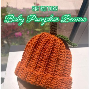 Op de afbeelding: Een gehaakte babymuts in oranje met een geribbelde textuur, voorzien van een bruine steel en een groen blaadje bovenop. De tekst PDF PATTERN Baby Pumpkin Beanie staat in groene letters boven de muts, die voor een raam wordt vastgehouden.
