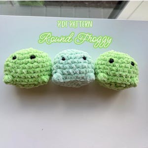 May include: Three round, plush, crocheted frog figures in shades of light green and mint. Each figure features two small black bead eyes. The text "PDF PATTERN Round Froggy" is displayed in light green lettering above the three crocheted items.