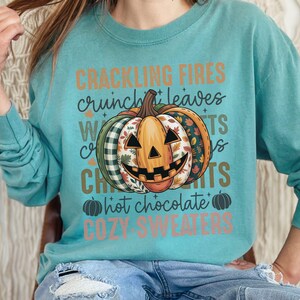 May include: A teal long-sleeved sweatshirt with a smiling jack-o'-lantern graphic and autumn patterns. The text on the shirt reads "Crackling Fires, Crunch Leaves, Witch Crafts, Hot Chocolate, Cozy Sweaters."