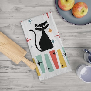 Puede incluir: Una toalla de cocina blanca con una ilustración de un gato negro, rayas de colores y detalles de estrellas. El gato mira hacia la derecha y tiene una estrella amarilla sobre su cabeza.