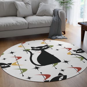 May include: A round white rug with a mid-century modern design featuring black cat silhouettes, starbursts, and atomic age-inspired shapes in red, yellow, and green. The rug is placed on a hardwood floor.