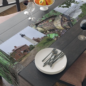 May include: A table runner with a collage of nature scenes. The runner features a lighthouse, a tree stump, and a view of the ocean. The runner is laid out on a dark wood table with a white plate and silverware.