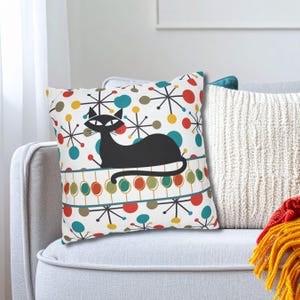 May include: A decorative throw pillow featuring a black cat design. The pillow has a white background with colorful atomic starburst and dot patterns in red, yellow, and teal. A teal and brown border runs across the bottom.