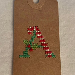 May include: A brown paper gift tag with a red, white, and green embroidered letter A. The letter is designed to look like a candy cane.