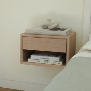 May include: A light wood floating nightstand with a drawer and a shelf. The shelf holds two books, one titled "Minimalista" and the other titled "Live Beautiful". A white ceramic sculpture sits on top of the nightstand.