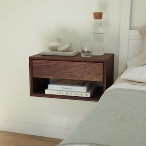 May include: A dark brown wooden floating nightstand with a drawer and a shelf. The shelf holds three books, one titled "Minimalista" and another titled "LIVE BEAUTIFUL". A clear glass bottle with a cork stopper and a clear glass cup are on top of the nightstand.