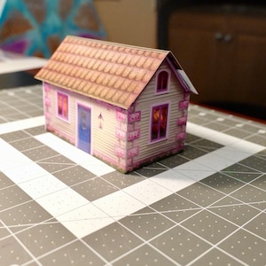 Op de afbeelding: Een klein papieren model van een huis met een bruin pannendak, roze muren en witte afwerking. Het huis heeft twee ramen en een deur.