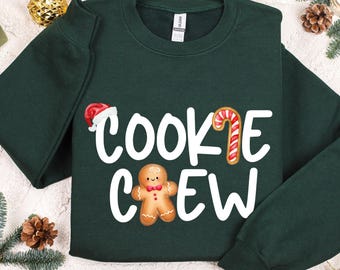 May include: A dark green sweatshirt with the words "COOKIE CREW" in white, featuring a Santa hat, candy cane, and gingerbread man graphic. Festive apparel for the holiday season.