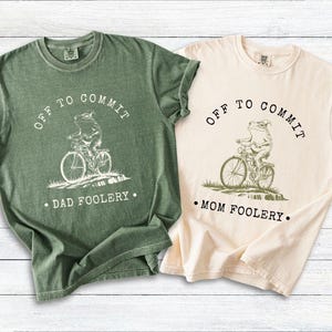 May include: Two t-shirts, one green and one cream-colored, with a whimsical frog riding a bicycle graphic. The green shirt reads "OFF TO COMMIT DAD FOOLERY." The cream shirt reads "OFF TO COMMIT MOM FOOLERY."