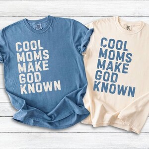 May include: Two short-sleeved t-shirts, one light blue and one cream-colored, are displayed. Each shirt features the phrase "COOL MOMS MAKE GOD KNOWN" in a distressed, blue font. The shirts are casual wear.