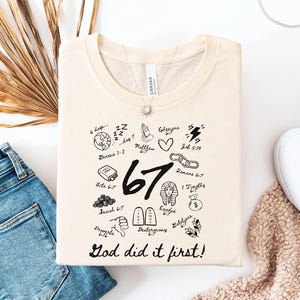 May include: Cream-colored t-shirt with a crew neck, featuring black graphic illustrations and text. The design includes the number "67", Bible verses, and the phrase "God did it first!" The shirt is folded, with blue jeans and white shoes in the background.