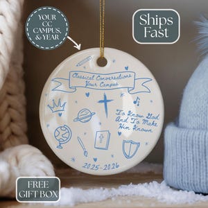 May include: A round, off-white ceramic ornament with blue illustrations and text. The ornament features a cross, crown, globe, and other symbols. Text includes "Classical Conversations Your Campus" and "To Know God And To Make Him Known."