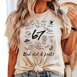 May include: Beige t-shirt with black graphic illustrations and the number 67. The shirt features the text "God did it first!" and various biblical references. The design includes a heart, lightning bolt, and other symbols.