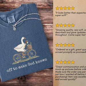 Puede incluir: Camiseta azul marino con una ilustraci&oacute;n de un ganso en bicicleta, con una cesta y una taza de caf&eacute;. El texto "off to make God known" est&aacute; impreso debajo. Un collar plateado est&aacute; sobre la camiseta.