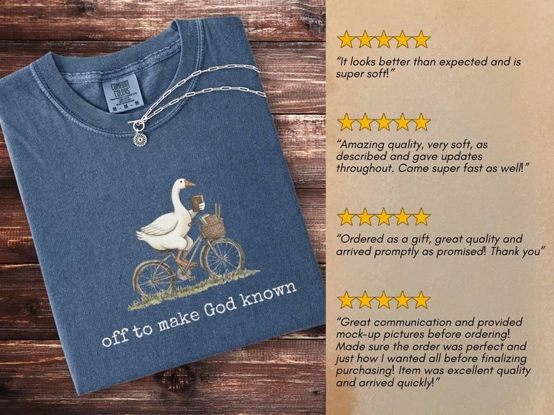 Puede incluir: Camiseta azul marino con una ilustraci&oacute;n de un ganso en bicicleta, con una cesta y una taza de caf&eacute;. El texto "off to make God known" est&aacute; impreso debajo. Un collar plateado est&aacute; sobre la camiseta.