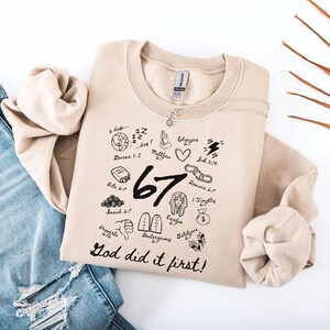 May include: Beige sweatshirt with black graphic illustrations and text. The design includes the number "67" and the phrase "God did it first!". A silver necklace is draped over the sweatshirt, which is paired with blue jeans.