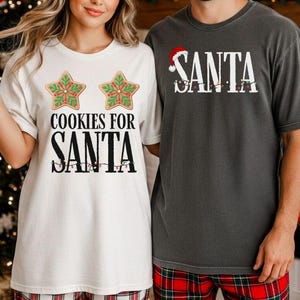 May include: Two matching Christmas-themed t-shirts. One is white with "COOKIES FOR SANTA" and star-shaped cookie graphics. The other is gray with "SANTA" and a Santa hat graphic. Both shirts have string light graphics.