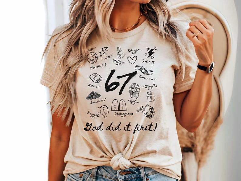 May include: Beige t-shirt with black graphic illustrations and the number 67. The shirt features the text "God did it first!" and various biblical references. The design includes a heart, lightning bolt, and other symbols.