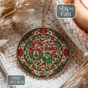 May include: Round Christmas ornament with a stained-glass design featuring the numbers "67" and the year "2025" in red and green. The ornament has a gold string for hanging and is decorated with poinsettias and holly leaves.