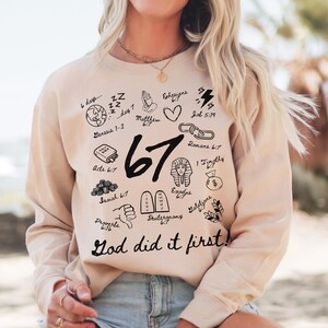 May include: A beige sweatshirt with black graphic illustrations and the text "67" and "God did it first!". The illustrations include a globe, a bible, a heart, and other religious symbols. The sweatshirt is a casual style.