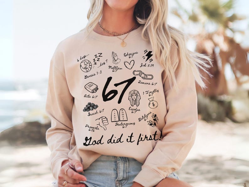 May include: A beige sweatshirt with black graphic illustrations and the text "67" and "God did it first!". The illustrations include a globe, a bible, a heart, and other religious symbols. The sweatshirt is a casual style.