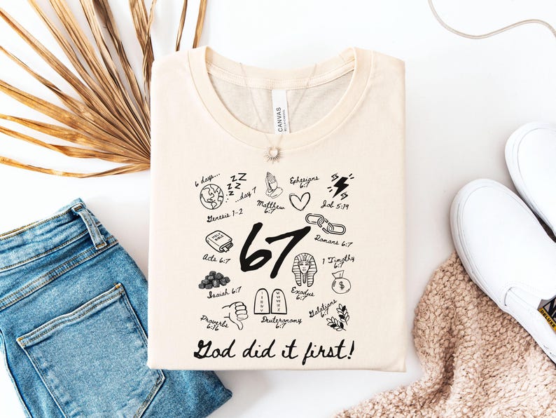 May include: Cream-colored t-shirt with a crew neck, featuring black graphic illustrations and text. The design includes the number "67", Bible verses, and the phrase "God did it first!" The shirt is folded, with blue jeans and white shoes in the background.