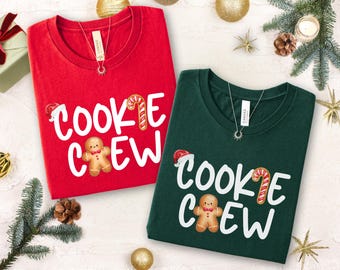 May include: Two crew neck t-shirts, one red and one green, with the words "COOKIE CREW" in white script. Each shirt features a gingerbread man, candy cane, and Santa hat graphic. Both shirts have a silver necklace.