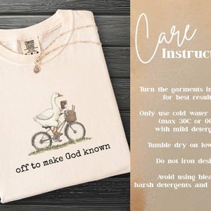 Puede incluir: Camiseta color crema con una ilustraci&oacute;n de un ganso en bicicleta y el texto "off to make God known". Tambi&eacute;n se ven las instrucciones de cuidado. La camiseta est&aacute; doblada sobre una superficie de madera oscura.