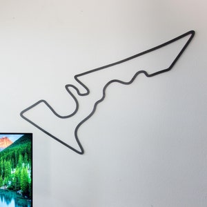 May include: A black metal outline of a race track is mounted on a white wall. A computer monitor displays a scenic landscape. A keyboard, mouse, and mousepad are on a brown desk.