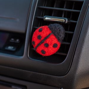 May include: A red and black ladybug car air vent clip. The ladybug is round with black spots and a black head. The clip is attached to a car's air vent, with a black dashboard and a red button visible.