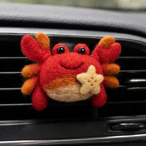 May include: A felt crab car air freshener in red, orange, and tan, with a smiling face and a starfish. The crab is attached to a car vent, with the radio and media controls visible below.