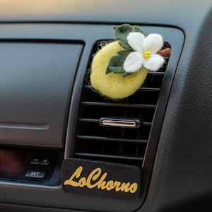 May include: A felt lemon car air freshener with a white flower and green leaves is inserted into a car vent. A black rectangular tag with the gold text "LoChorno" is below the vent.