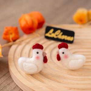 May include: Two handmade white felt chicken figurines with red combs and waddles, and pink cheeks, sit on a wooden tray. A black tag with the text "LaChorva" in yellow is behind the chickens. Orange fruit is in the background.