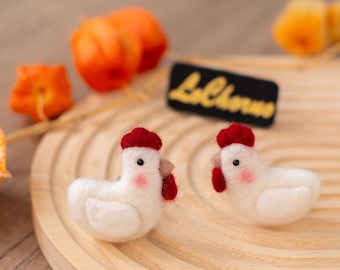May include: Two handmade white felt chicken figurines with red combs and waddles, and pink cheeks, sit on a wooden tray. A black tag with the text "LaChorva" in yellow is behind the chickens. Orange fruit is in the background.