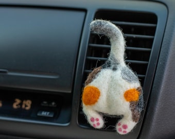 May include: A felted cat butt car air vent accessory. The cat's rear is white with brown, gray, and orange markings. The tail is gray, and the paws are pink. The accessory is inserted into a car's air vent.