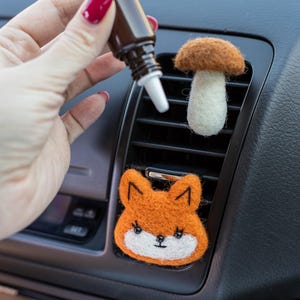 May include: Orange fox and mushroom car air fresheners. The fox has a white face and black details. The mushroom has a white stem and a brown cap. A brown bottle with a dropper is held nearby.
