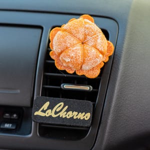 May include: An orange felt air freshener shaped like a citrus fruit, placed in a car vent. Below the vent is a black rectangle with the gold text "LoChorno". The car's dashboard is black.