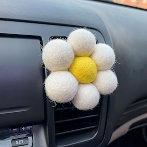 May include: A white felt flower car air freshener with a yellow center. The flower is attached to a car's air vent.