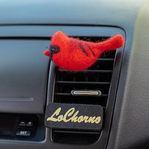 May include: A bright red needle-felted cardinal bird ornament with black facial markings and a tail, attached to a rectangular black felt base featuring the gold embroidered text LoChorno, designed to clip onto a vehicle air vent.