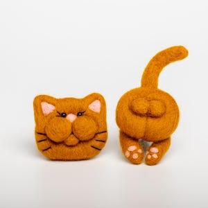 May include: Two orange felt cat figures. One is a cat face with pink ears and nose, black whiskers, and black eyes. The other is a cat rear with a tail and pink paw pads. The figures are on a white background.