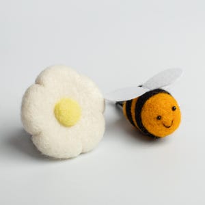 May include: A felted bee with orange and black stripes and white wings, next to a white felt flower with a yellow center. The bee has a smiling face. The items are on a white background.