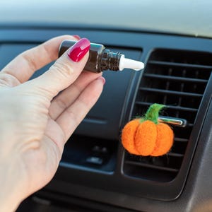May include: An orange felt pumpkin car air freshener with a green stem is clipped to a car vent. A brown bottle with a white dropper is held nearby, suggesting the use of essential oils for fragrance.