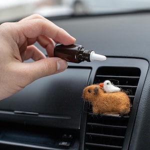 May include: A brown essential oil bottle with a white dropper is held above a felted car air vent diffuser. The diffuser is a brown guinea pig with a white bird on its back. The car's dashboard is black.