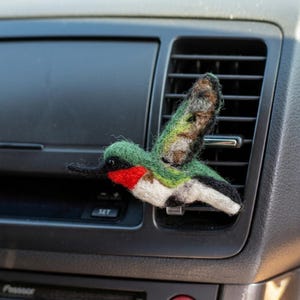 May include: A needle-felted hummingbird ornament featuring green, white, and black feathers with a bright red throat patch. The bird has a long, thin black beak and a small black eye, and it is attached to a clear plastic clip for mounting.