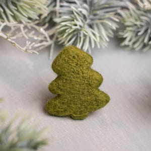May include: A handcrafted, olive green felt Christmas tree ornament. The tree is a simple, stylized design with rounded edges. The background features frosted evergreen branches, suggesting a winter holiday theme.