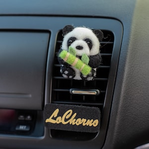 May include: A felt panda air freshener with black and white fur, holding a green bamboo stalk. The panda is attached to a car vent. A black rectangular tag with the word "LoChorno" in gold script is below the vent.