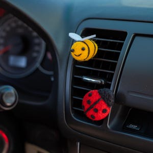 May include: A yellow and black felt bee and a red and black felt ladybug hanging from the air vent of a car.