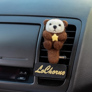 May include: A brown and white felt otter car air vent clip holding a yellow star. The clip is attached to a black vent. A black rectangular tag with the gold text "LoChorno" is below the otter.