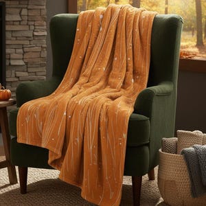 May include: An orange throw blanket with white linear and dot patterns draped over a green armchair. The scene includes a fireplace, a small wooden table with a mug and pumpkin, and a basket with blankets, creating a cosy autumn atmosphere.