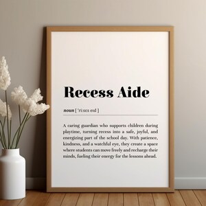 May include: A framed print with the words "Recess Aide" in a bold, black font. The definition of "Recess Aide" is below the title. A white vase with white flowers is to the left of the print.